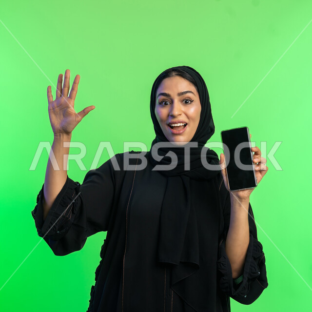 Mobile phone applications and services display, gestures of happiness and pleasure, use of modern technical devices, close-up portrait of a veiled Saudi Arabian Gulf woman holding a mobile phone with a blank black screen, a young Emirati woman raising her hand with expressions of peace and surprise, green background, chroma key