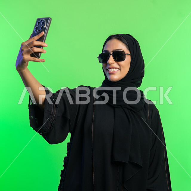 توثيق اللحظات السعيدة على وسائل التواصل الاجتماعي، استخدام الأجهزة التقنية الحديثة،  بورتريه مقرب لامرأة عربية خليجية سعودية محجبة ترتدي عباءة سوداء تلتقاط صور سيلفي تذكارية عبر الهاتف، شابة اماراتية تنظر الى الموبايل بإيماءات السعادة، خلفية خضراء، كروما