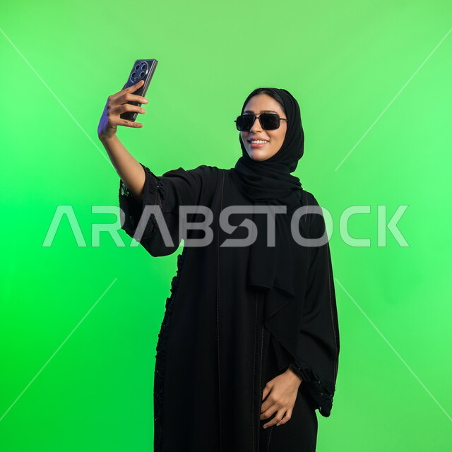 استخدام الأجهزة التقنية الحديثة، توثيق اللحظات السعيدة على وسائل التواصل الاجتماعي، بورتريه لامرأة عربية خليجية سعودية محجبة ترتدي عباءة سوداء تلتقاط صور سيلفي تذكارية عبر الهاتف، شابة اماراتية تنظر الى الموبايل بإيماءات السعادة، خلفية خضراء، كروما