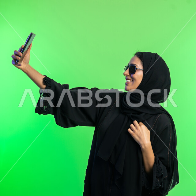 مشاركة اللحظات السعيدة على وسائل التواصل الاجتماعي، استخدام الأجهزة التقنية الحديثة،  بورتريه مقرب لامرأة عربية خليجية سعودية محجبة ترتدي عباءة سوداء تلتقاط صور سيلفي تذكارية عبر الهاتف، شابة اماراتية تنظر الى الموبايل بإيماءات السعادة، خلفية خضراء، كروما