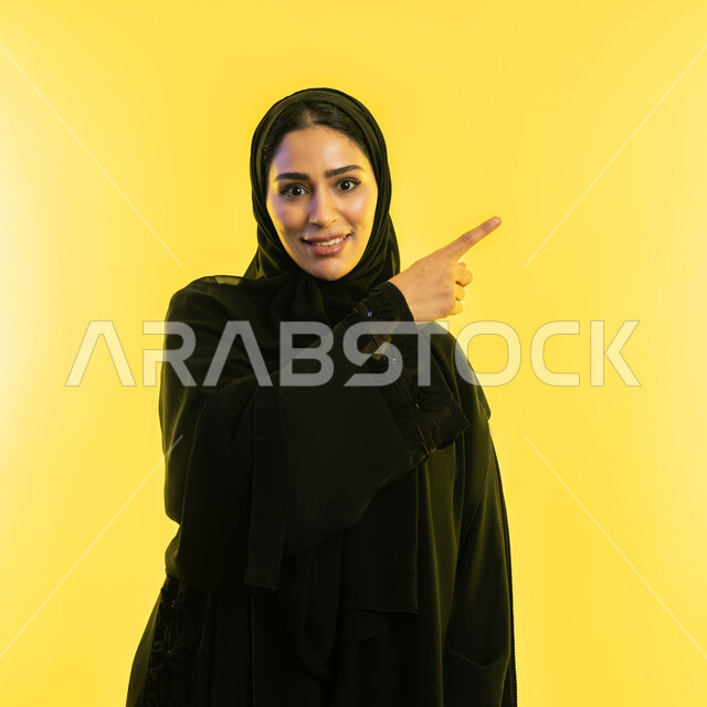 حركات وإشارات يدين تدل على هذا المكان (هنا)، تحديد موقع معين بإيماءات البشاشة، شابة اماراتية تنظر الى الكاميرا بإيماءات السرور، بورتريه مقرب لامرأة عربيه خليجيه سعوديه مبتسمه محجبه ترتدي العباءة السوداء ترفع اصبها السبابة تشير الى الاعلى، خلفية صفراء