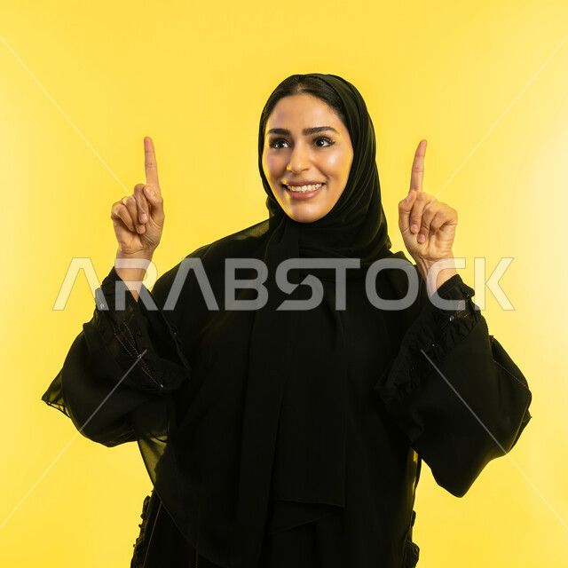تحديد موقع معين بإيماءات البشاشة، شابة اماراتية تنظر الى شيء ما بتعابير السرور، إشارات وحركات يدين تدل على هذا المكان (هنا)، بورتريه مقرب لامرأة عربيه خليجيه سعوديه مبتسمه محجبه ترتدي العباءة السوداء ترفع اصابعها وتشير الى الأعلى، خلفية صفراء