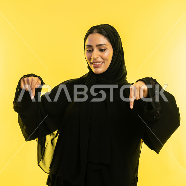 إشارات وحركات يدين تدل على هذا المكان (هنا)، تحديد موقع شيء معين، بورتريه مقرب لامراة عربية خليجية اماراتية مبتسمة محجبة ترتدي العباءة السوداء تشير بإصبعي السبابة إلى جهة الأسفل، شابة سعودية تقف باستقامة، خلفية صفراء