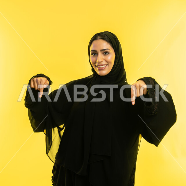 تحديد موقع شيء معين، إشارات وحركات يدين تدل على هذا المكان (هنا)، بورتريه مقرب لامراة عربية خليجية اماراتية مبتسمة محجبة ترتدي العباءة السوداء تشير بإصبعي السبابة إلى جهة الأسفل، شابة سعودية تقف باستقامة وتنظر الى الكاميرا، خلفية صفراء