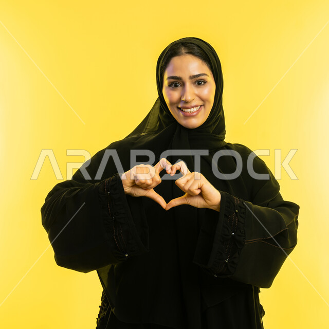 Showing feelings of love and passion, concept of love and affection, heart shaped hand gestures, close-up portrait of veiled Saudi Arabian Gulf woman wearing black abaya, young Emirati woman looking at camera with expressions of pleasure and happiness, yellow background