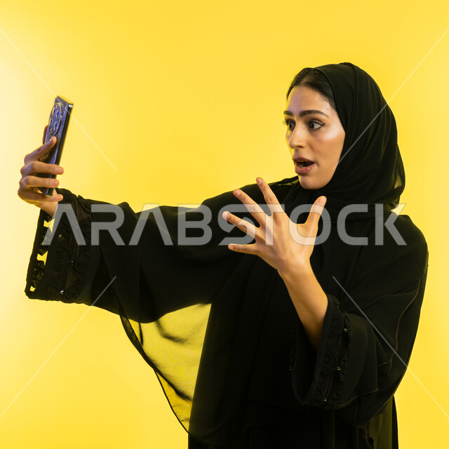 ايماءات الاندماج والسرور، بث مباشر ودردشة ومحادثات مع الأقارب عن بعد، بورتريه مقرب لإمرأة عربية خليجية سعودية محجبة ترتدي عباءة سوداء تتواصل مع الاصدقاء عبر الهاتف المحمول، شابة اماراتية تقومبإجراء مكالمات الفيديو، خلفية صفراء