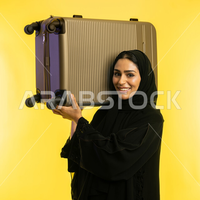 إجازة الصيف السياحية, الشعور بالسعادة والفرح، الاستعداد لقضاء رحلة صيفية ترفيهية ممتعة, بورتريه مقرب لشابة عربية خليجية سعودية محجبة ترتدي عباءة سوداء ترفع حقيبة السفرعلى كتفها، امرأة اماراتية تنظر إلى الكاميرا بإيماءات الحماس, خلفية صفراء
