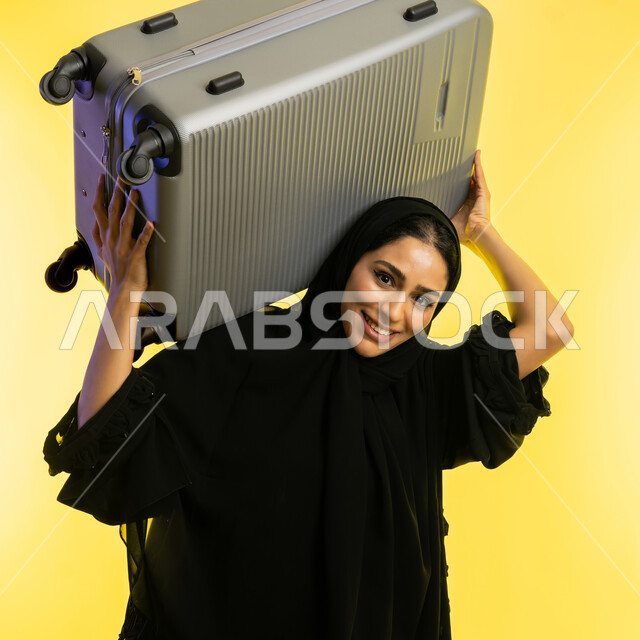 الاستعداد لقضاء رحلة صيفية ترفيهية ممتعة, السياحة في إجازة الصيف, بورتريه مقرب لشابة عربية خليجية سعودية محجبة ترتدي عباءة سوداء تحمل حقيبة السفر على كتفها، امرأة اماراتية تنظر إلى الكاميرا بإيماءات الحماس, الشعور بالسعادة والفرح، خلفية صفراء