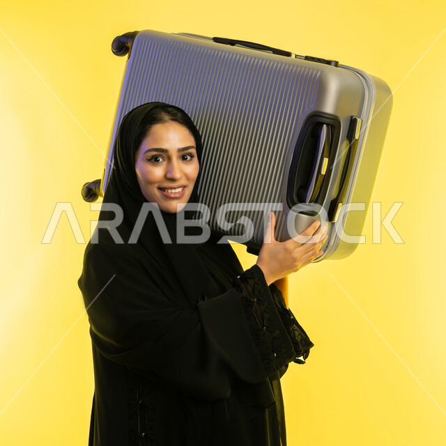 السياحة في إجازة الصيف, الاستعداد لقضاء رحلة صيفية ترفيهية ممتعة, بورتريه مقرب لشابة عربية خليجية سعودية محجبة ترتدي عباءة سوداء تحمل حقيبة السفر على كتفها، امرأة اماراتية تنظر إلى الكاميرا بإيماءات الحماس, الشعور بالسعادة والفرح، خلفية صفراء