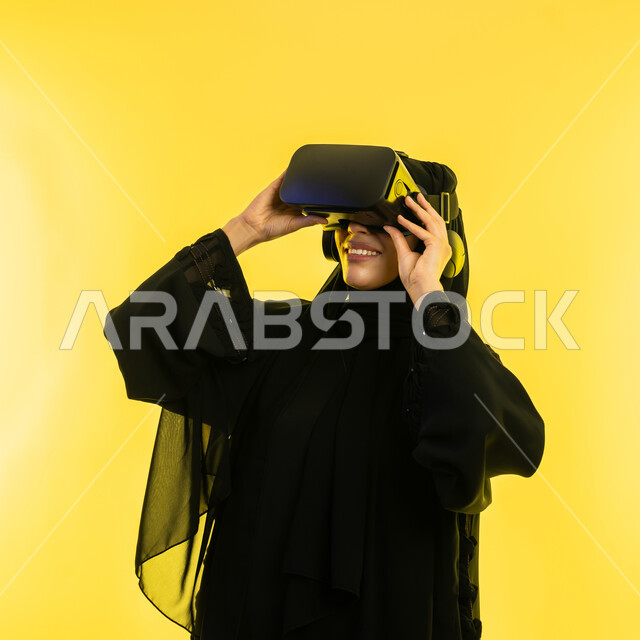 Using modern electronic technologies, gestures of interaction and integration with holographic elements, close-up portrait of a veiled Saudi Arabian Gulf woman wearing a black abaya and 3D virtual reality (VR) glasses, a young Emirati woman watching favorite movies and series, yellow background