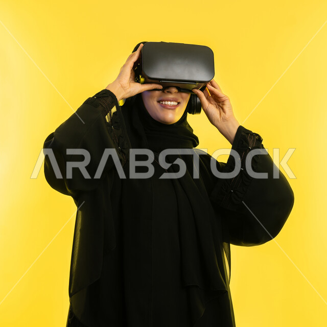استخدام التقنيات الإلكترونية الحديثة، إيماءات التفاعل والاندماج مع العناصر الهولوجرامية، بورتريه مقرب لامرأة عربية خليجية سعودية محجبة ترتدي عباءة سوداء ونظارات الواقع الإفتراضي (في آر) ثلاثية الأبعاد، شابة اماراتية تشاهد الأفلام والمسلسلات المفضلة، خلفية صفراء