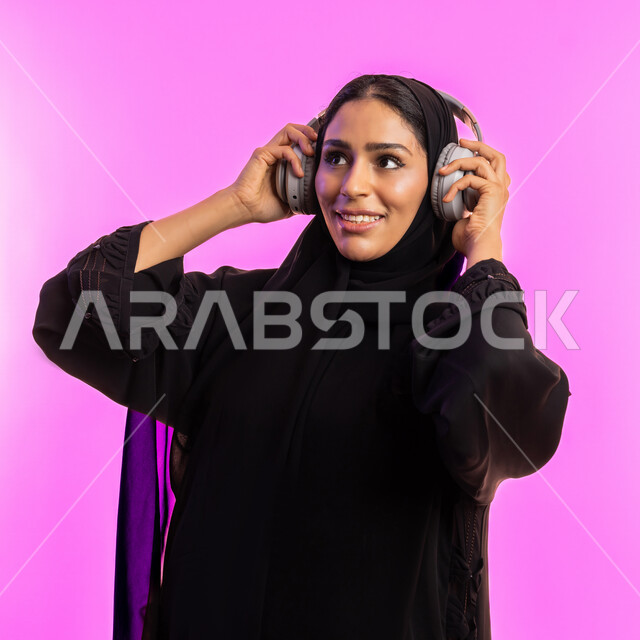 إستخدام تقنيات الهيدفون، الاستمتاع في أوقات الفراغ, مفهوم الإصغاء والإستماع، بورتريه مقرب لسيدة عربية خليجية سعودية مبتسمة محجبة ترتدي عباءة سوداء تضع سماعات الرأس الصوتية، امرأة اماراتية تستمع إلى الموسيقى بإيماءات السعادة والاندماج، خلفية وردية