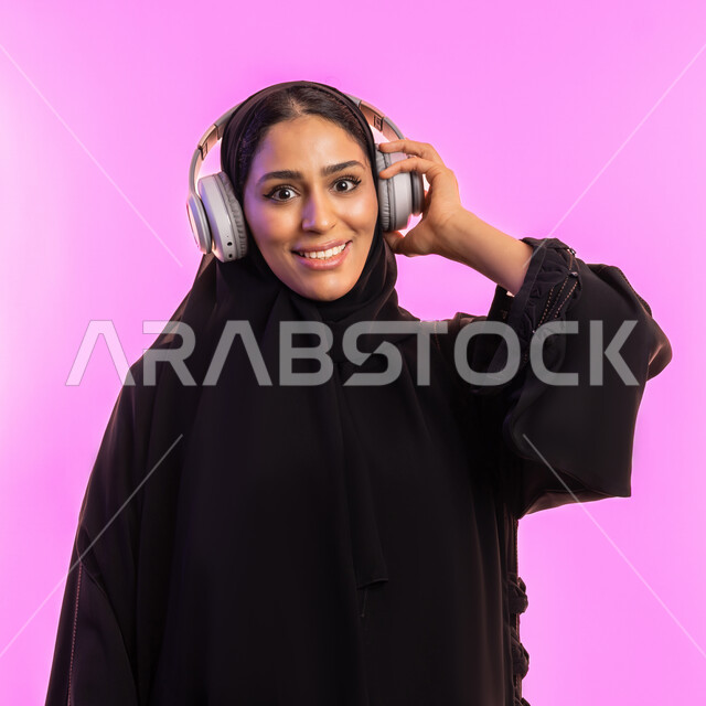 الاستمتاع في أوقات الفراغ, مفهوم الإصغاء والإستماع، إستخدام تقنيات الهيدفون، بورتريه مقرب لسيدة عربية خليجية سعودية مبتسمة محجبة ترتدي عباءة سوداء تضع سماعات الرأس الصوتية، امرأة اماراتية تستمع إلى الموسيقى بإيماءات السعادة والاندماج، خلفية وردية