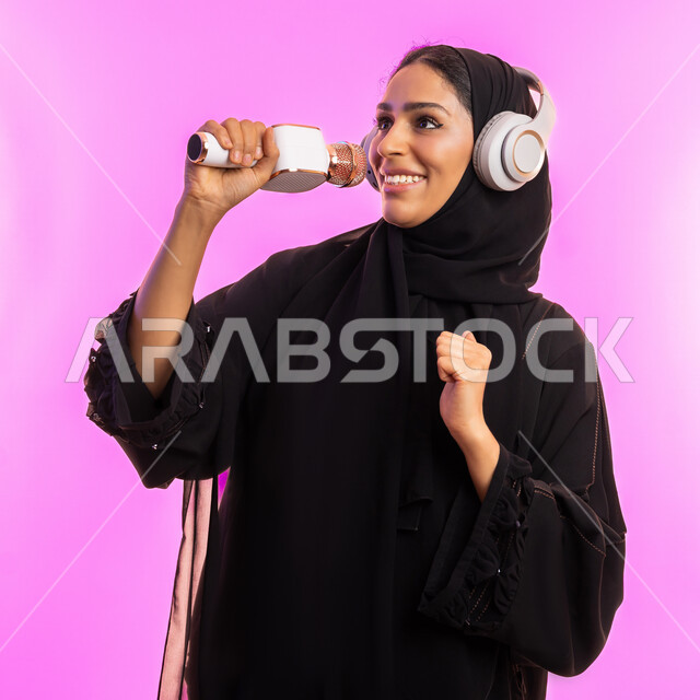 The concept of art and its positive impact on women, starting vocal training, having a good time performing vocal performances, close-up portrait of a veiled Saudi Arabian Gulf young woman wearing a black abaya holding a microphone and wearing headphones, Emirati woman practicing singing talent with gestures of pleasure, pink background