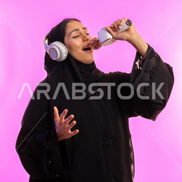 قضاء وقت ممتع بأداء العروض الصوتية، بدء التدريب الصوتي، مفهوم الفن وتأثيره الايجابي على الامرأة، بورتريه مقرب لشابة عربية خليجية سعودية محجبة ترتدي عباءة سوداء تمسك المايكروفون وتضع سماعات الرأس، سيدة اماراتية تمارس موهبة الغناء بإيماءات السرور، خلفيه وردية