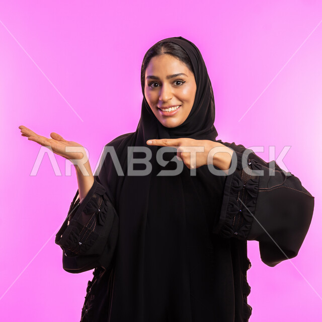 ايماءات التأكد وتحديد مكان معين, الوقوف باستقامة والاشارة إلى شيء ما، سيدة اماراتية تنظر إلى الكاميرا بتعابير السعادة والسرور, بورتريه مقرب لامرأة عربيه خليجيه سعودية مبتسمة ترتدي حجاب والعباءة السوداء ترفع اصبعها السبابة للاختيار, خلفية وردية