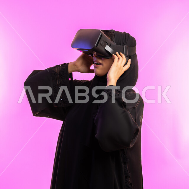 Gestures of interaction and integration with holographic elements, concept of modern electronic technologies, close-up portrait of a veiled Saudi Arabian Gulf woman wearing a black abaya and 3D virtual reality (VR) glasses, young Emirati woman watching favorite movies and series, pink background