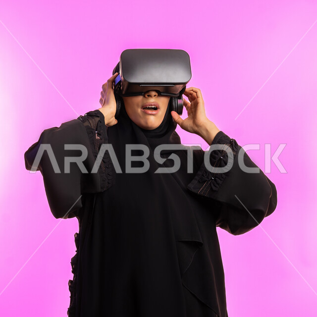مفهوم التقنيات الإلكترونية الحديثة، إيماءات التفاعل والاندماج مع العناصر الهولوجرامية، بورتريه مقرب لامرأة عربية خليجية سعودية محجبة ترتدي عباءة سوداء ونظارات الواقع الإفتراضي (في آر) ثلاثية الأبعاد، شابة اماراتية تشاهد الأفلام والمسلسلات المفضلة، خلفية وردية