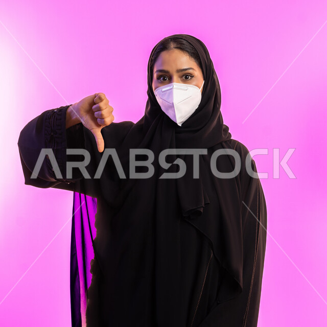 الإنزعاج من شيء ما، الالتزام بالاحترازات الوقائيه ضد كوفيد 19، امرأة عربية خليجية سعودية ترتدي حجاب وعباءة سوداء تشير بإصبع الإبهام للأسفل بإيماءات عدم الموافقة والرفض، بورتريه مقرب لشابة إماراتية ترتدي الكمامة تنظر إلى الكاميرا، خلفية وردية