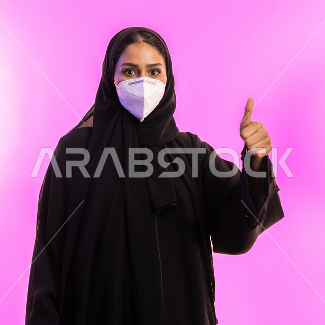الحفاظ على السلامة و تجنب انتشار العدوى بكوفيد 19، إجراء الاحترازات الوقائية من فايروس كورونا، شابة اماراتية تنظر الى الكاميرا بايماءات القبول والرضا, بورتريه مقرب لإمرأة عربية سعودية خليجية محجبة ترتدي الكمامة ترفع اصبع الإبهام بعلامة الجودة, خلفية وردية