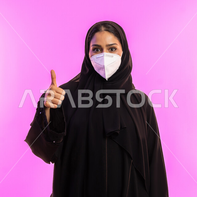 إجراء الاحترازات الوقائية من فايروس كورونا، شابة اماراتية تنظر الى الكاميرا بايماءات القبول والرضا, الحفاظ على السلامة و تجنب انتشار العدوى بكوفيد 19، بورتريه مقرب لإمرأة عربية سعودية خليجية محجبة ترتدي الكمامة ترفع اصبع الإبهام بعلامة الجودة, خلفية وردية