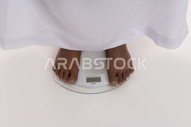 الإلتزام بحمية غذائية صحية، إيجاد حل للمشاكل الصحية، خسارة الدهون والسعرات الحرارية، بورتريه مقرب لرجل عربي خليجي اماراتي ممتلئ يقف على الميزان لقياس وزنه، المعاناة من زيادة والوزن والسمنة المفرطة، خلفية بيضاء
