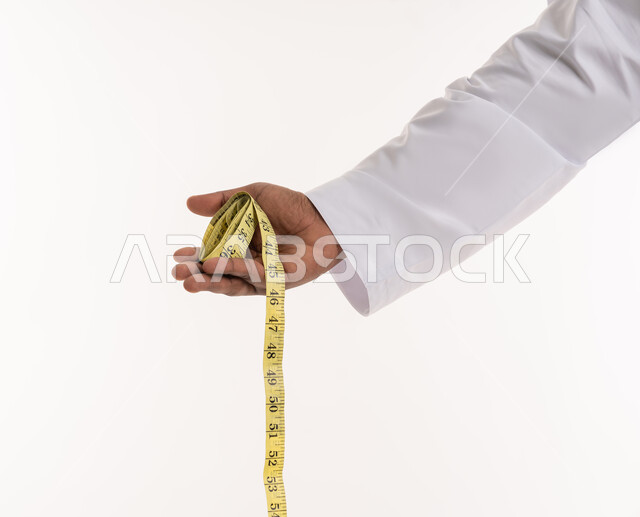 الإلتزام بنادي للتمارين الرياضية، خسارة السعرات الحرارية، المشاكل الصحية والمعاناة من الوزن الزائد، إتباع حمية غذائية صحية، بورتريه مقرب ليد رجل عربي خليجي اماراتي يحمل بيده شريط مقياس الخصر، خلفية بيضاء