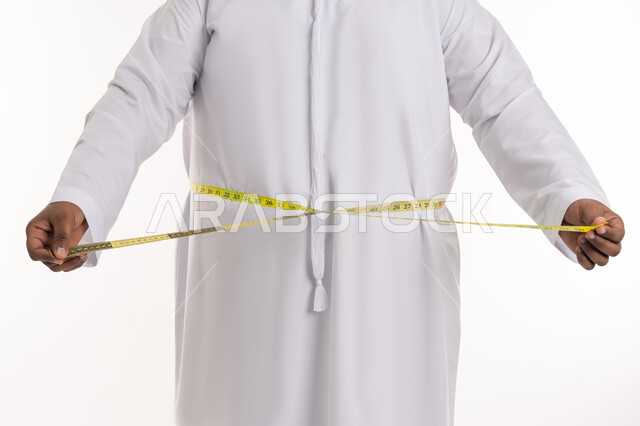 إتباع حمية غذائية صحية، المعاناة من الوزن الزائد والمشاكل الصحية، بورتريه مقرب لرجل عربي خليجي اماراتي ممتلئ يرتدي الكندورة الاماراتية يقوم بقياس محيط خصره عبر الشريط المقياس، الإلتزام بنادي للتمارين الرياضية، خسارة السعرات الحرارية، خلفية بيضاء