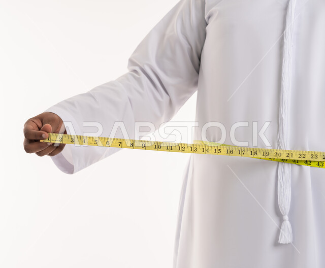 المعاناة من الوزن الزائد والمشاكل الصحية، بورتريه مقرب لرجل عربي خليجي اماراتي ممتلئ يرتدي الكندورة الاماراتية يقوم بقياس محيط خصره عبر الشريط المقياس، إتباع حمية غذائية صحية، الإلتزام بنادي للتمارين الرياضية، خسارة السعرات الحرارية، خلفية بيضاء