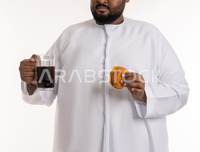 ضرر الإكثار من أكل الوجبات السريعة، طلب واستلام الأطعمة الخارجية، بورتريه مقرب لرجل عربي خليجي اماراتي يرتدي الكندورة الاماراتية يحمل بيده قطعة من البرجر اللذيذ ومشروب غازي، تناول الطعام الغير الصحي، مطاعم للمأكولات الغربية في الامارات، خلفية بيضاء