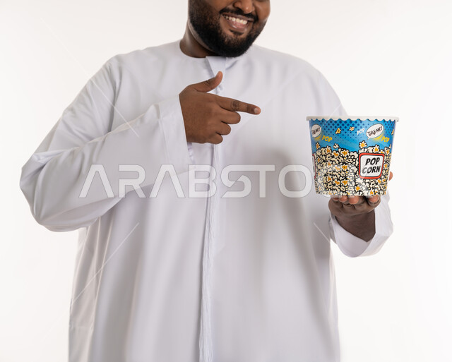 انشطة ترفيهية منزلية، تناول الوجبات الخفيفة والسناك، بورتريه مقرب لرجل عربي خليجي اماراتي يرتدي الكندورة الاماراتية يشير بيده الى علبة من الفوشار المقرمش، الاستمتاع بمشاهدة الأفلام والمسلسلات، متابعة برامج التلفاز، قضاء وقت ممتع في المنزل، خلفية بيضاء