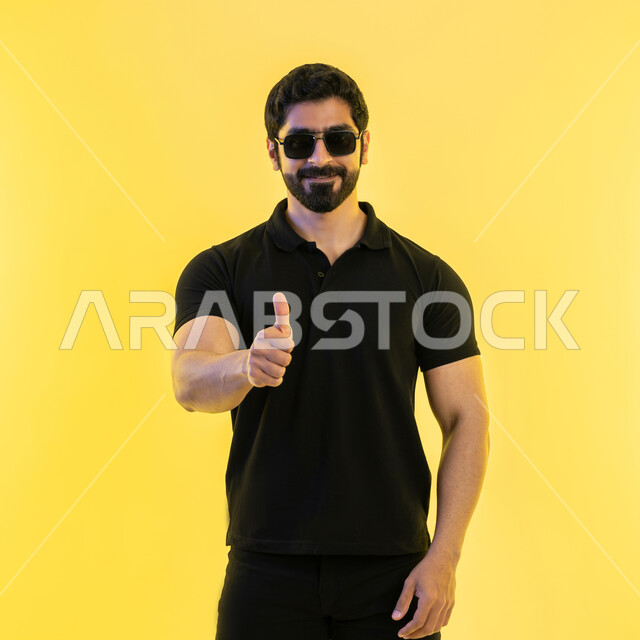 التعبير عن الجودة والاتقان، الموافقة والرضا و القبول، الوقوف بسرور وبشاشة وسعادة، بورتريه لشاب عربي خليجي إماراتي يرتدي الزي الكاجوال يرفع ابهامه بإيماءات الجودة والامتياز والتميز، خلفية صفراء