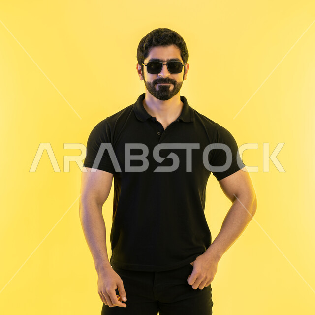 مفهوم اللياقة والرشاقة، المحافظة على التوازن الجسدي، امتلاك جسم رياضي صحي، الأناقة والاهتمام بالمظهر الخارجي، بورتريه لشاب عربي خليجي اماراتي مبتسم يرتدي الزي الكاجوال والنظارة الشمسية ينظر إلى الكاميرا بإيماءات الثقة بالنفس، الشعور بالسرور والبهجة، خلفية صفراء