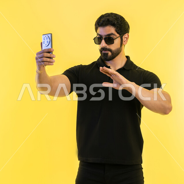 اجراء مكالمة فيديو عبر تطبيقات ومواقع السوشال ميديا، التقاط صور سيلفي تذكارية عبر الجوال، استخدام الأجهزة والتقنيات الحديثة، بورتريه لشاب عربي خليجي اماراتي يرتدي الزي الكاجوال يقوم بحمل الهاتف المحمول، خلفية صفراء