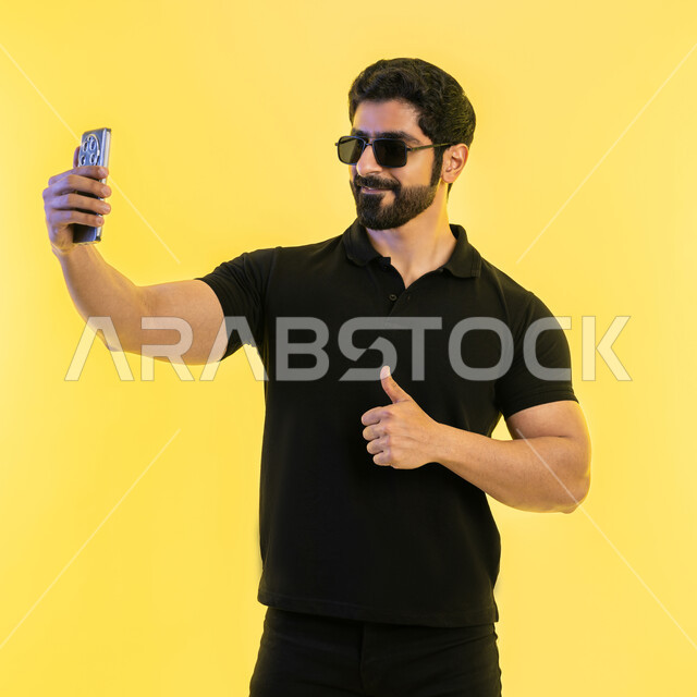 التقاط صور سيلفي تذكارية عبر الجوال، اجراء مكالمة فيديو عبر تطبيقات ومواقع السوشال ميديا، استخدام الأجهزة والتقنيات الحديثة، بورتريه لشاب عربي خليجي اماراتي يرتدي الزي الكاجوال والنظارة الشمسية يرفع اصبع الابهام إيماءات الجودة والامتياز والتميز، خلفية صفراء