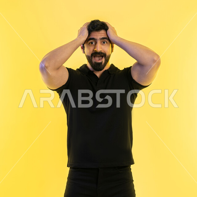 الوقوف بإيماءات التعجب والدهشة، التفاجؤ والاستغراب من شيء ما، بورتريه لشاب عربي خليجي إماراتي يرتدي الزي الكاجوال يضع يديه على رأسه بتعابير الصدمة والذهول، الانبهار وعدم تصديق أمر معين، خلفية صفراء