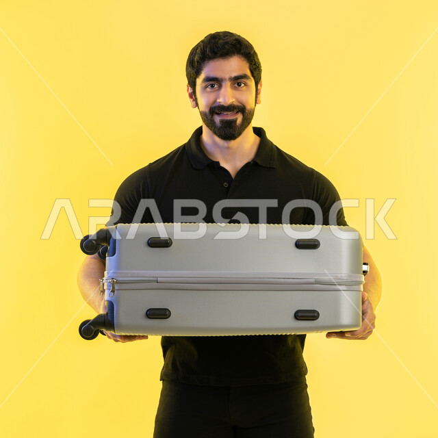 الاستعداد لقضاء رحلة صيفية ترفيهية ممتعة، السياحة في إجازة الصيف، بورتريه مقرب لشاب عربي خليجي اماراتي يرتدي الزي الكاجوال يحمل حقيبة السفر، النظر إلى الكاميرا بإيماءات الحماس، الشعور بالسعادة والفرح، خلفية صفراء