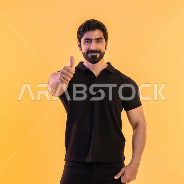 الوقوف بسرور وبشاشة وسعادة، الموافقة والرضا و القبول، التعبير عن الجودة والاتقان، بورتريه لشاب عربي خليجي إماراتي يرتدي الزي الكاجوال يرفع ابهامه بإيماءات الجودة والامتياز والتميز، خلفية صفراء