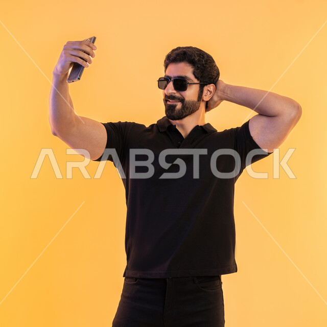 Taking commemorative selfies via mobile, making a video call via social media applications and sites, using modern devices and technologies, portrait of a young Arab Gulf Emirati man wearing casual clothes and sunglasses holding a mobile phone, yellow background
