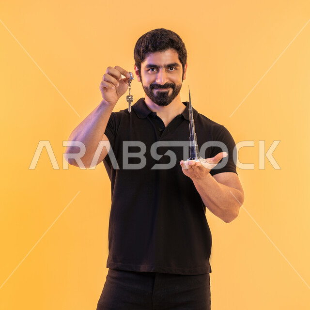 النمو والتطور العمراني للابراج وناطحات السحاب، مفهوم امتلاك شقة جديدة، الفخر بمعالم الامارات العربية المتحدة، بورتريه لشاب عربي اماراتي خليجي مبتسم يرتدي الزي الكاجوال يحمل بيده مجسم ثلاثي الابعاد لبرج خليفة الشهير ومفتاح، خلفية صفراء