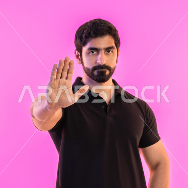 Standing in anger and refusing a specific order, being silent and stopping talking, asking not to interfere and lowering the voice and threatening, refraining from doing a specific order, close-up portrait of a young Arab Gulf Emirati man wearing casual clothes raising his hand up in gestures of annoyance, pink background