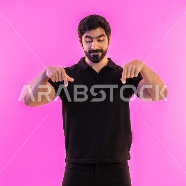 حركات تدل على هذا المكان (هنا)، البشاشة والسرور وتحديد موقع معين، بورتريه لشاب عربي خليجي اماراتي مبتسم يرتدي الزي الكاجوال يشير بأصابع السبابة نحو الأسفل، تأكيد حدوث شيء هذه اللحظة، خلفية وردية