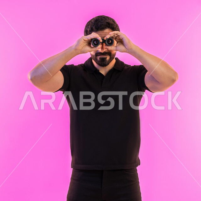 رؤية الاشياء البعيدة عبر التلسكوب، التخطيط المستقبلي للأهداف والطموحات، بورتريه لشاب عربي خليجي اماراتي يرتدي الزي الكاجوال ينظر من خلال المنظار ثنائي العينين، حب المغامرة والاستكشاف، خلفية وردية