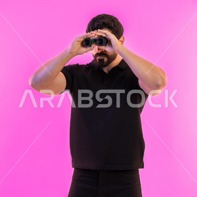 التخطيط المستقبلي للأهداف والطموحات، بورتريه لشاب عربي خليجي اماراتي يرتدي الزي الكاجوال ينظر من خلال المنظار ثنائي العينين، رؤية الاشياء البعيدة عبر التلسكوب، حب المغامرة والاستكشاف، خلفية وردية