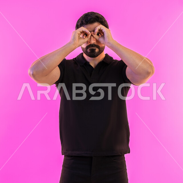 رؤية الاشياء البعيدة، حب المغامرة والاستكشاف، التخطيط المستقبلي للأهداف والطموحات، بورتريه لشاب عربي خليجي اماراتي يرتدي الزي الكاجوال يضع يديه على عينه على شكل منظار ثنائي العينين، خلفية وردية