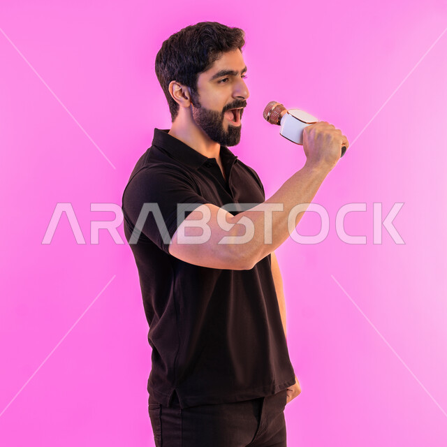 Performing stage performances, practicing singing talent, enjoying vocal talent performance, side portrait of a young Arab Gulf Emirati man wearing casual clothes holding a microphone, gestures and expressions of integration and interaction with music, recording a singing audio clip, pink background