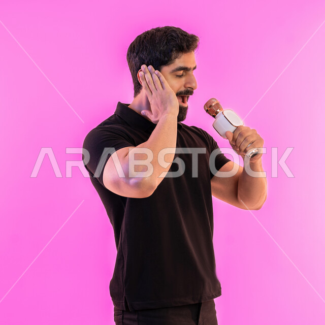 الاستمتاع بأداء المواهب الصوتية، تقديم العروضات المسرحية، ممارسة موهبة الغناء، بورتريه من الجانب لشاب عربي خليجي اماراتي يرتدي الزي الكاجوال يمسك بيده المايكروفون، إيماءات وتعابير الاندماج والتفاعل مع الموسيقا، تسجيل مقطع صوتي غنائي، خلفية وردية
