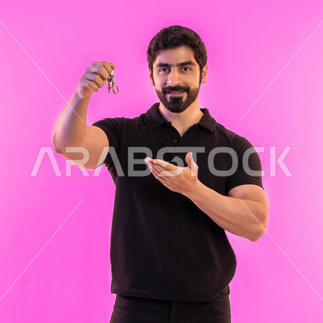 اقتناء سيارة جديدة بإيماءات السرور، الحصول على رخصة قيادة، بورتريه لشاب عربي خليجي اماراي مبتسم يرتدي الزي الكاجوال يشير بيده إلى مفتاح السيارة، خدمة صف السيارة بالمواقف الخاصة، استلام وتسليم المفاتيح، خلفيه وردية
