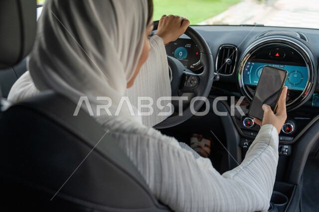 Freedom to drive for Arab women, an Emirati Gulf Arab woman who tracks the route online, women’s self-reliance in transportation, a young Saudi woman sitting inside her car holding a mobile phone, the concept of independence and self-reliance, driving on public roads and streets