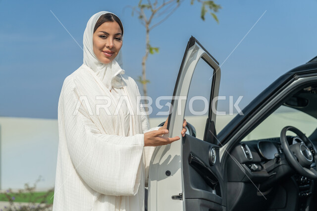 مفهوم الاعتماد على النفس عند الأنثى الخليجية, حرية قيادة السيارة للمرأة العربية, شابة عربية خليجية إماراتية تلبس العباءة البيضاء تقف بجانب سيارتها, سيدة سعودية تفتح باب السيارة وتشير إليه بإيماءات السعادة, مفهوم الاستقلال عند السيدات العربيات